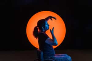 A performer holds a sideways sitting yoga pose on the background of a lit-up orange circle and a black surrounding.