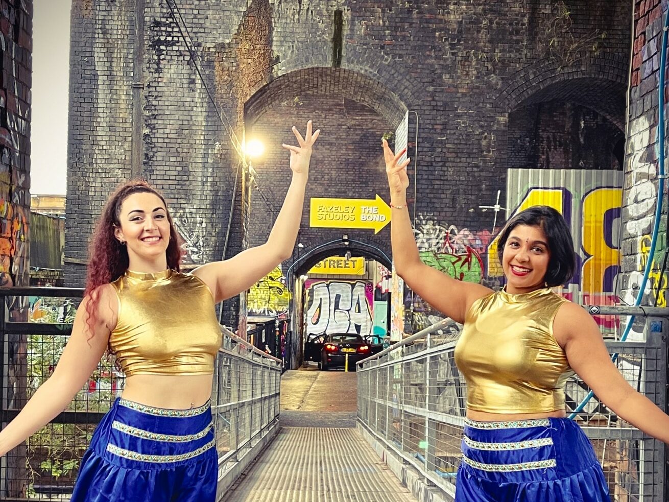 Two smiling dancers in Bollywood costumes pose in a tunnel.