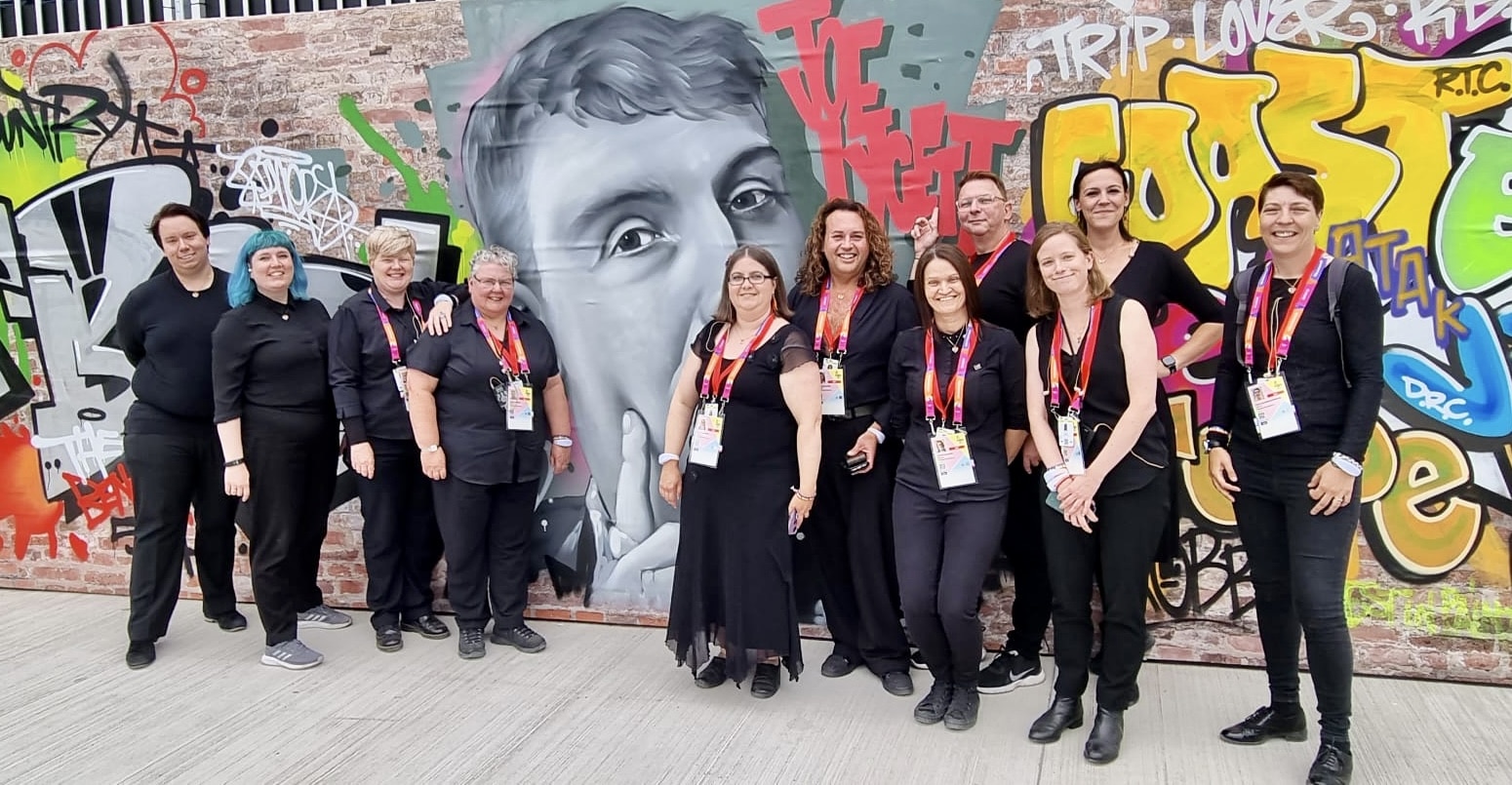 Rainbow voices choir - A group of choir members pose in front of a colourful graffiti wall.