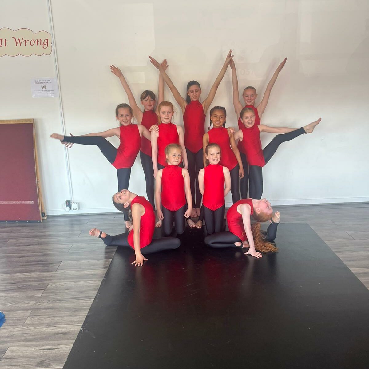Performers dressed in red colorful suits pose in a dance formation in a practice room.