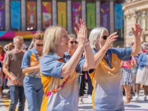 Two volunteers in orange and blue uniforms dance in Victoria Square during the Birmingham 2022 Commonwealth Games.
