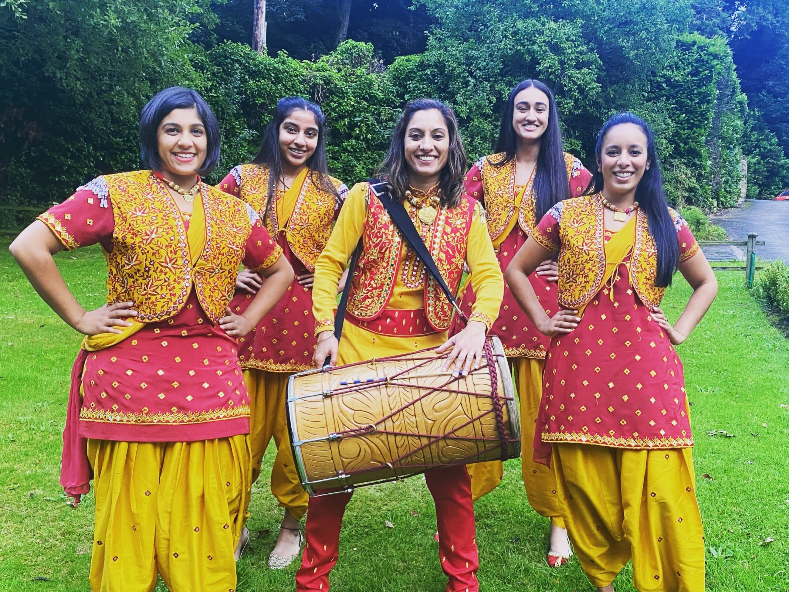 Five women in traditional Indian costumes pose with a drumming instrument.