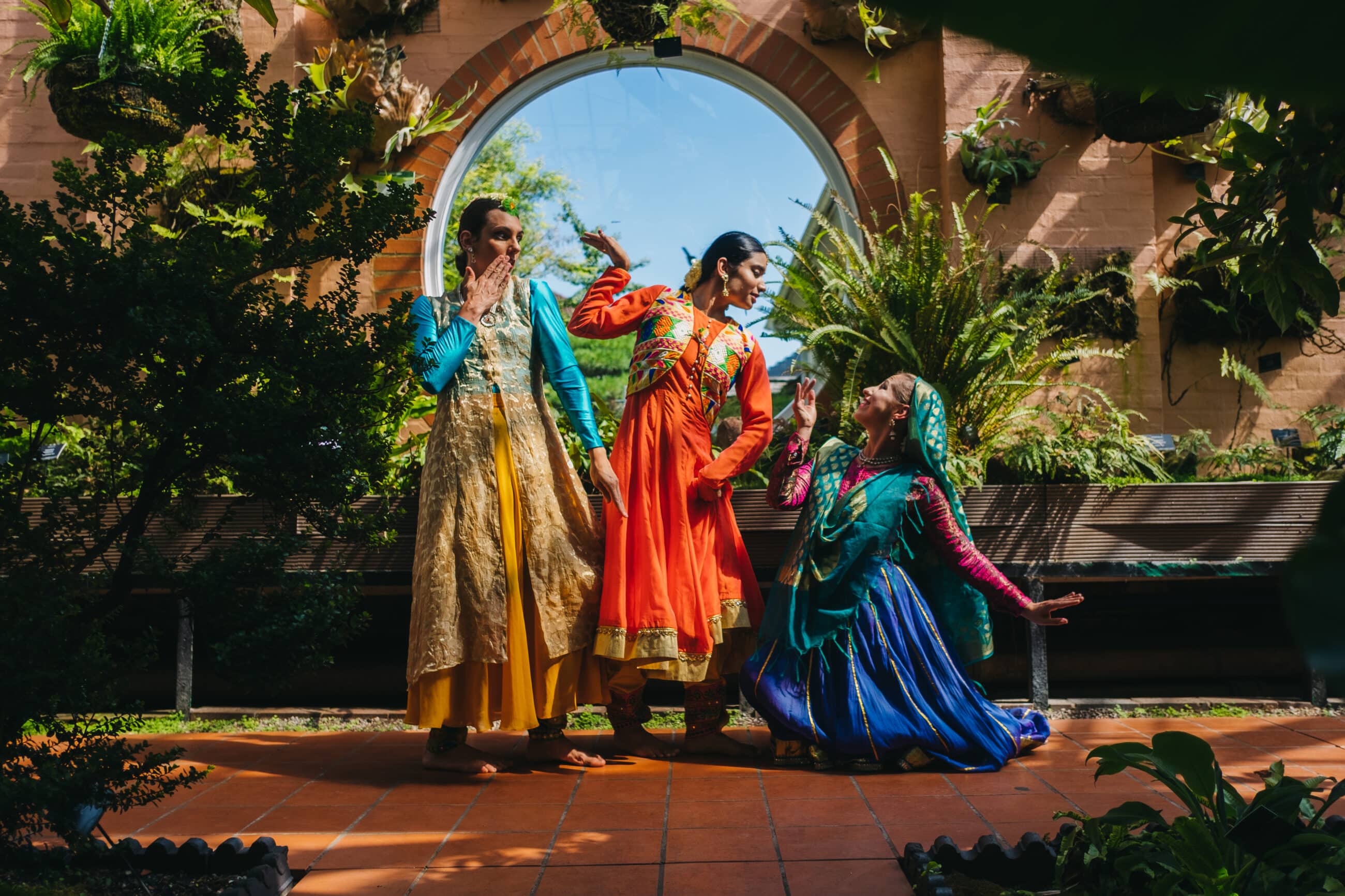 Three performers in traditional Persian costumes pose front of a brick and greenery wall.