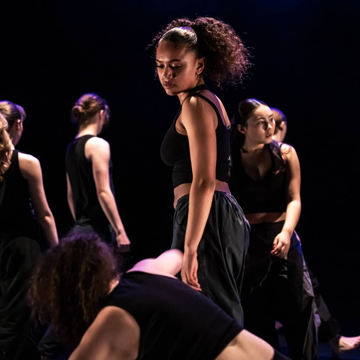 Six young dancers perform a pensive piece in a dim lit background.