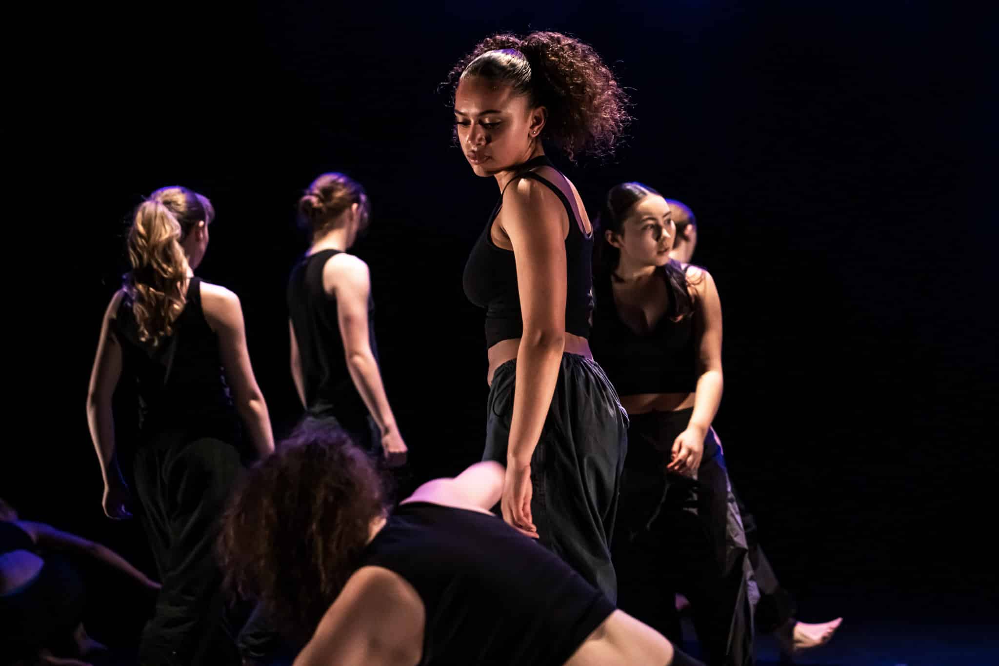 Six young dancers perform a pensive piece in a dim lit background.
