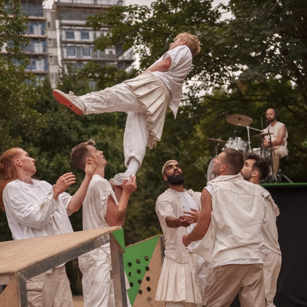 Five dancers dressed in neutral clothing throw and catch another dancer in an outdoor background.