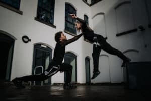 A male dancer kneels on the ground and holds a female dancer who's jumping towards him.