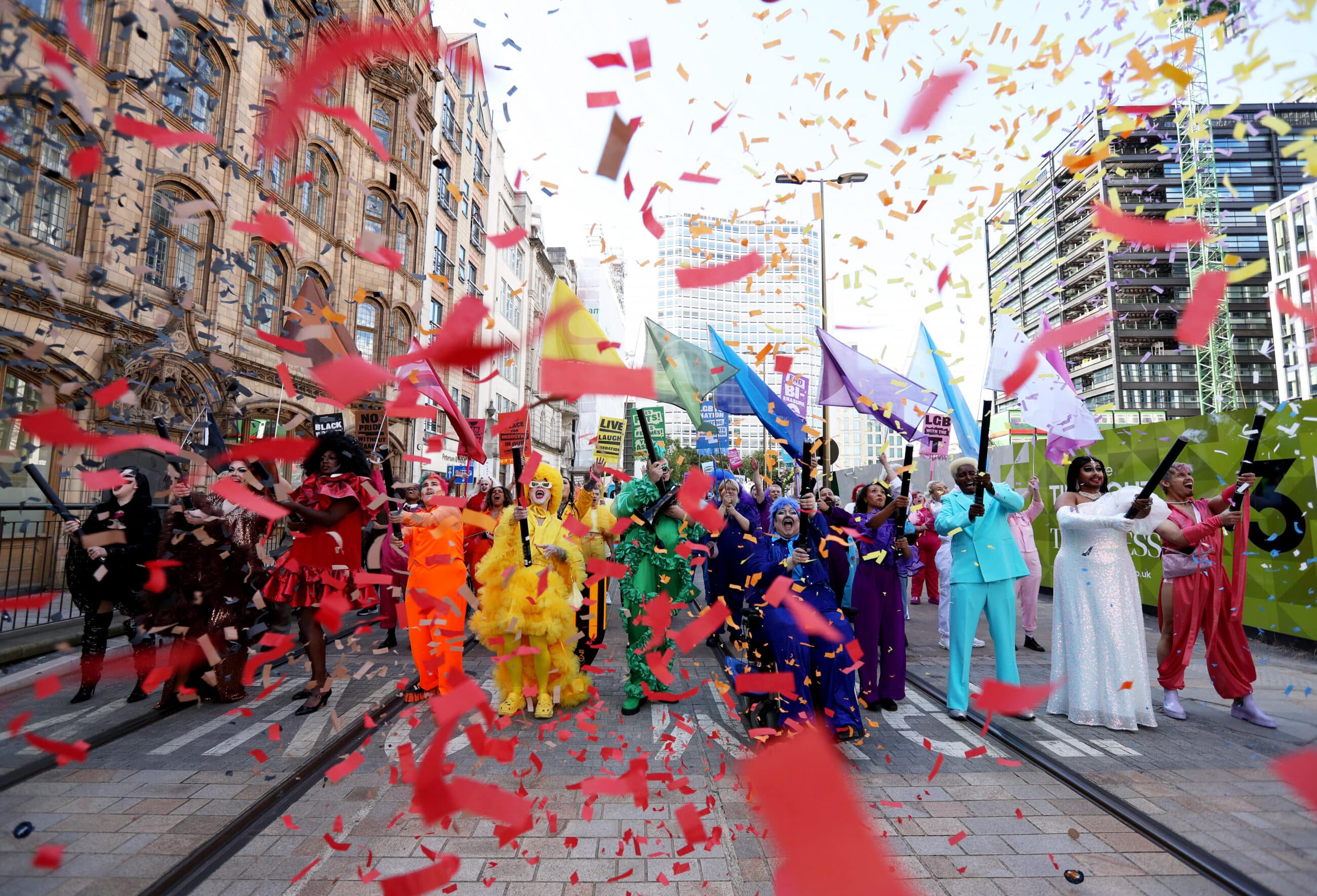 Performers dressed in bright rainbow clothing and waving colored rainbow flags march down the street while confetti rains down in the frame.