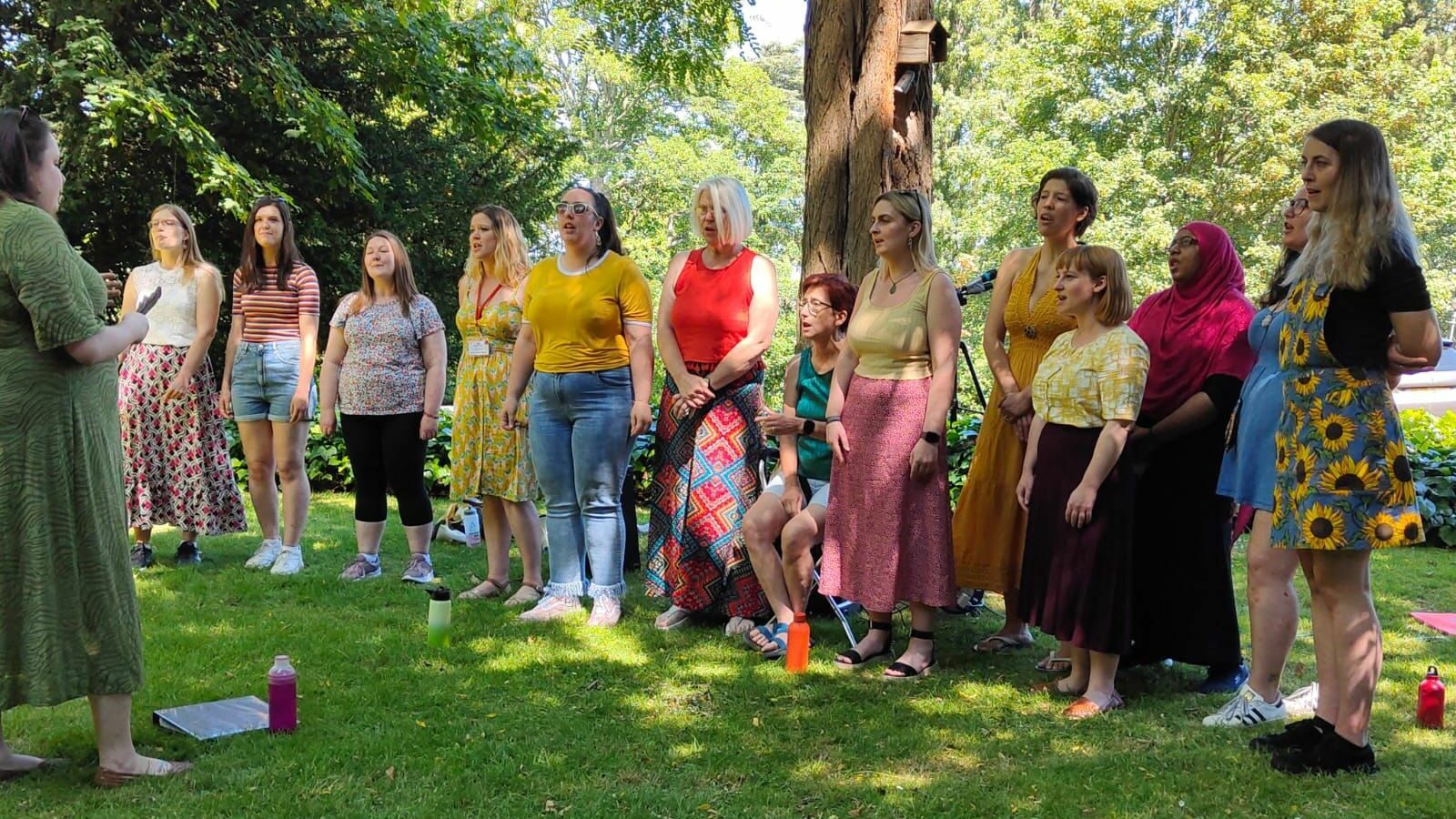 Female choir members dressed in colorful clothing sing in a park, led by an instructor.