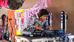 A woman focuses on playing a DJ deck on a colorful background.