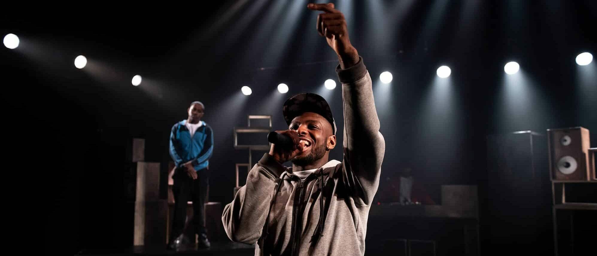 A performer sings into the mic to the background of a dim-lit stage, smiling, holding his hand up.