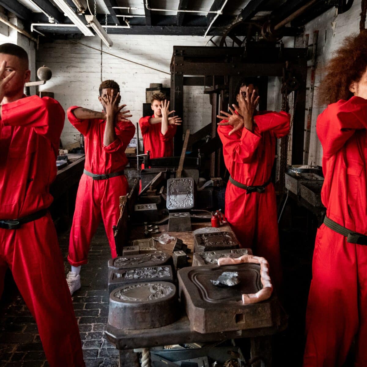 Five performers dressed in red pose with their hands crossed in a factory room.