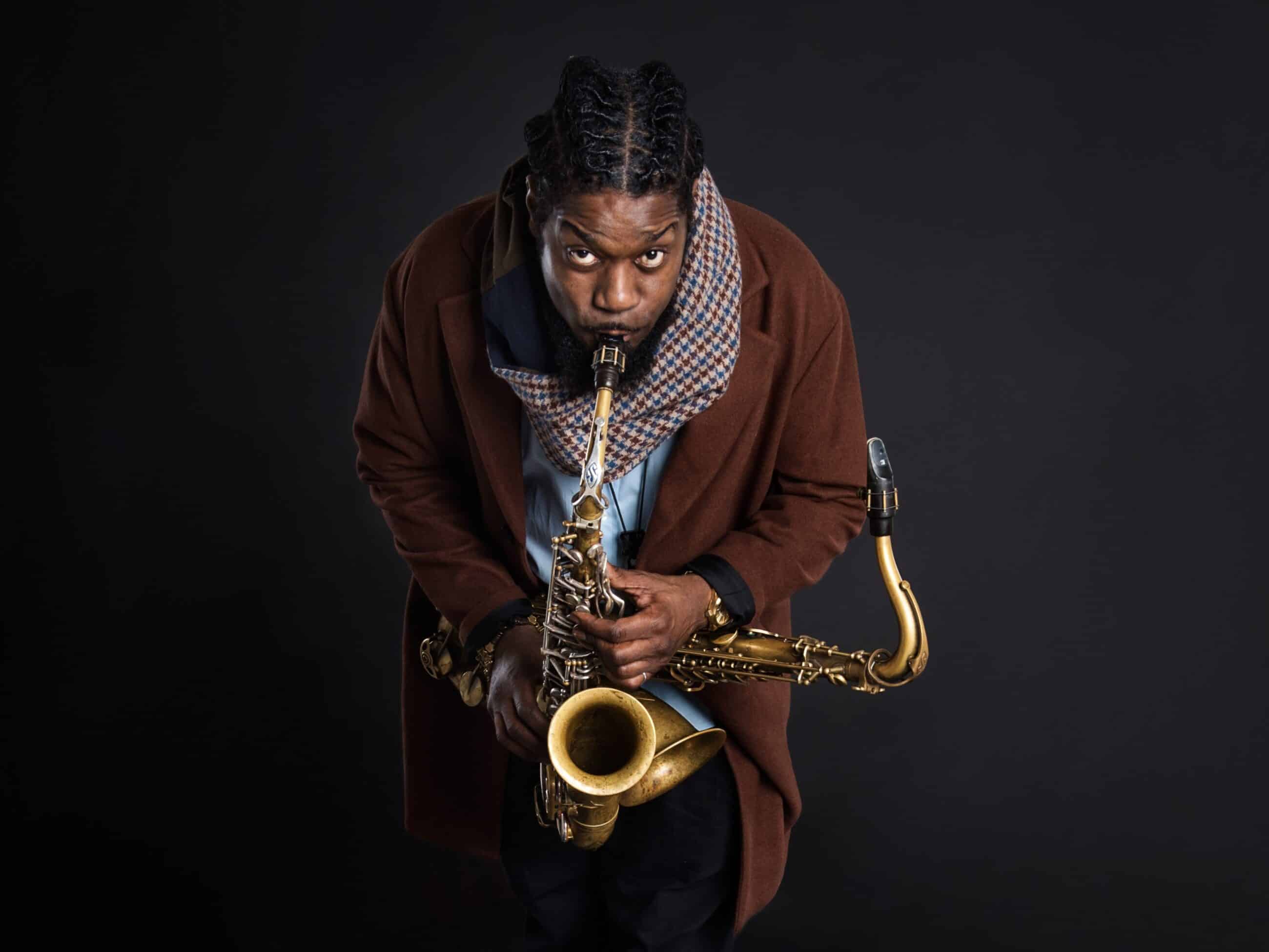 Soweto Kinch poses with his saxophone on a black background.