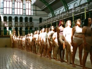 Women dressed in nude clothing stand in a row on the background of a repurposed bath house.