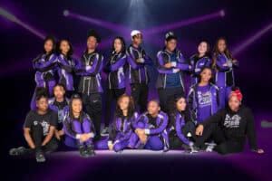 Young dancers dressed in purple team costumes pose on a dark background.