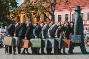 A cast of street performers dressed in old-fashioned suits and carrying suitcases lay in line on a statue.